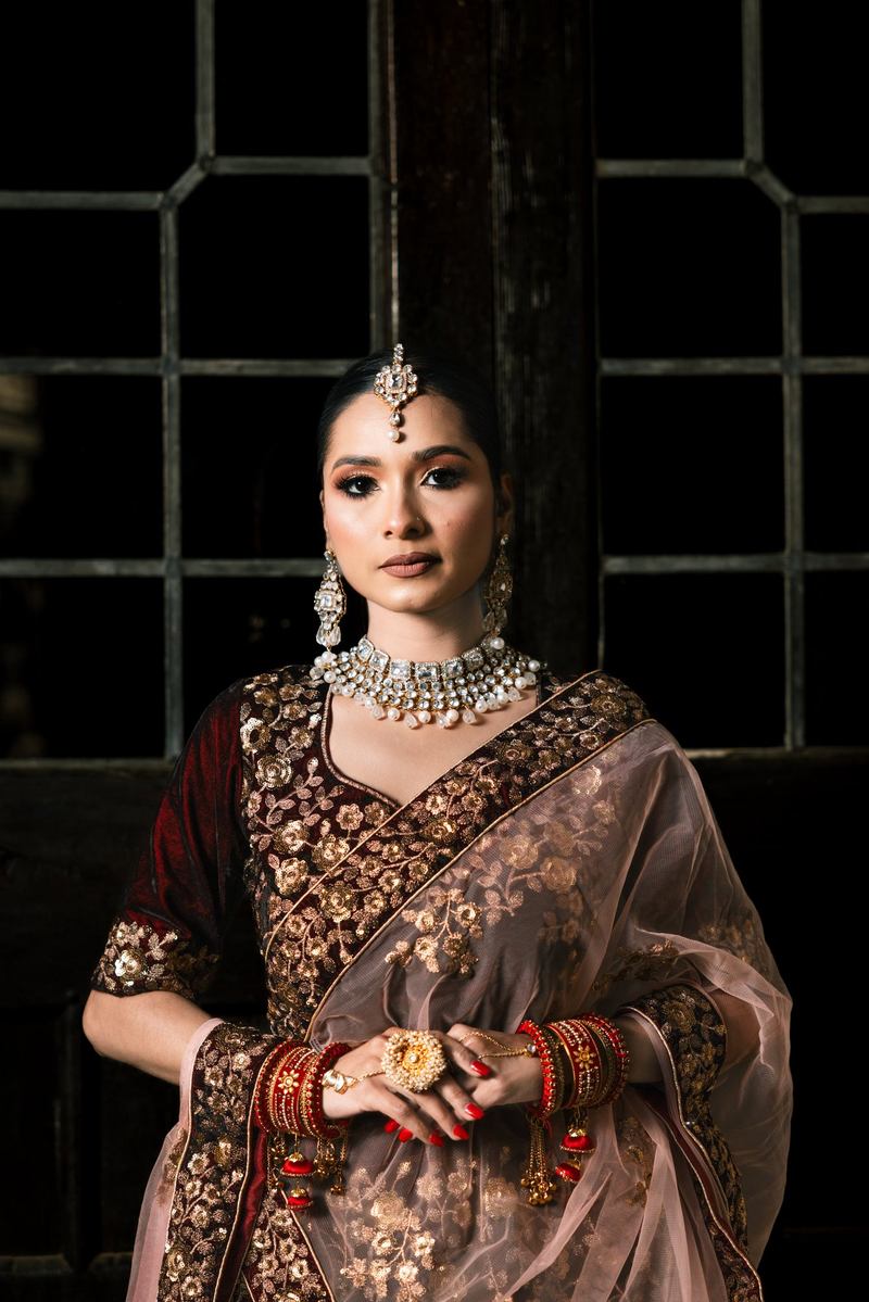 Bride in maroon and gold with kundan jewelry, hands clasped, looking directly at the camera before dark windows