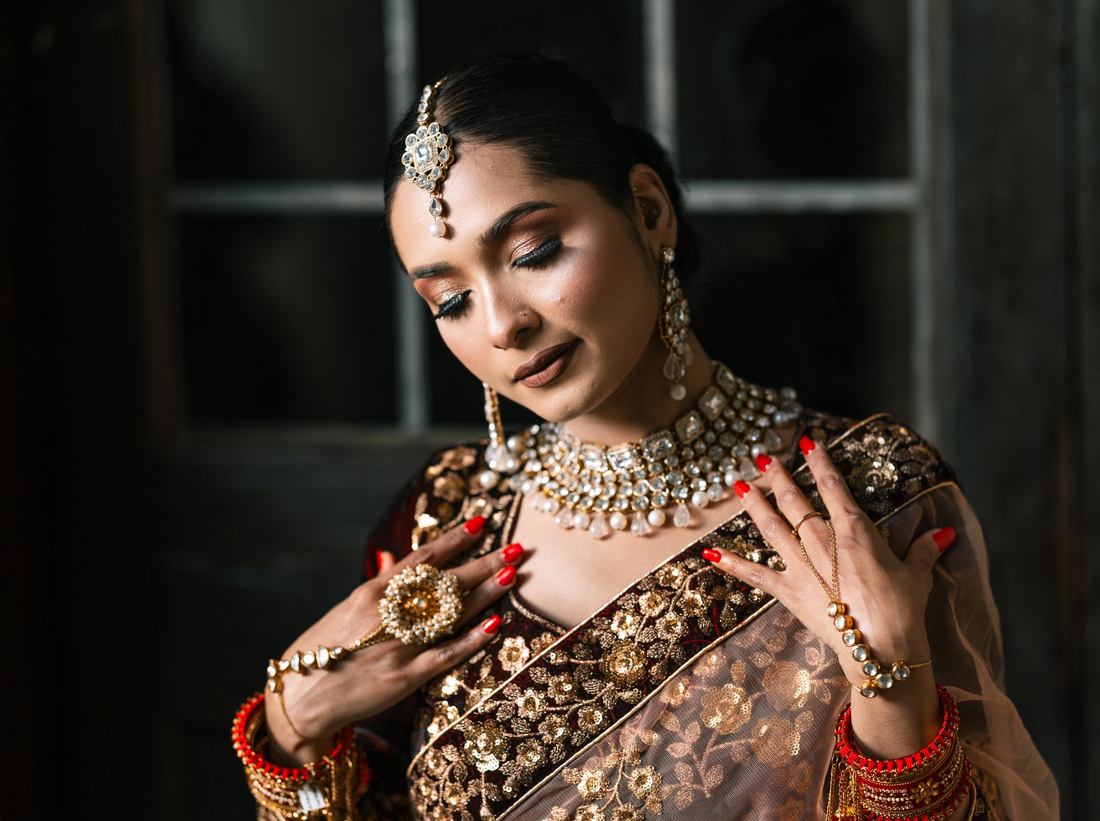 South Asian bride with eyes closed and hands resting at her kundan necklace, in front of a dark paned window