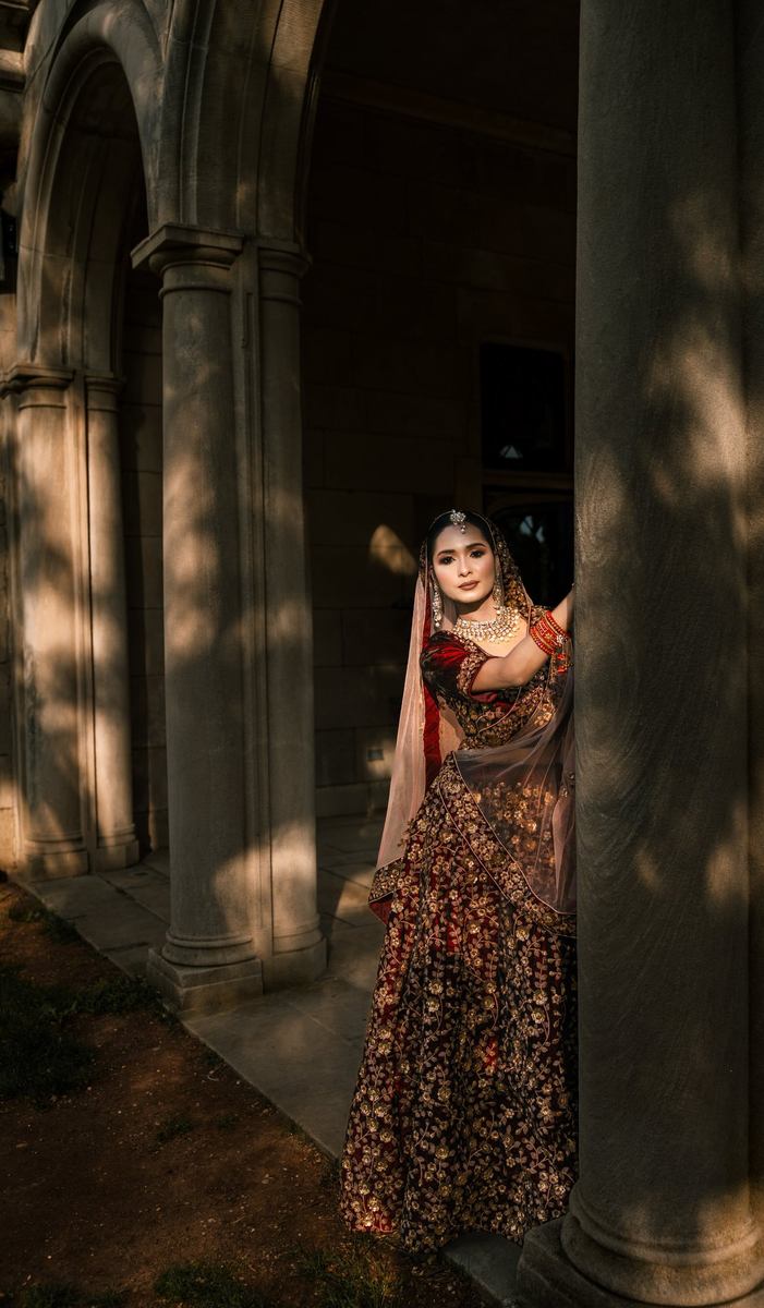 Bride in a maroon embroidered lehenga standing between tall stone columns in warm, dappled evening light