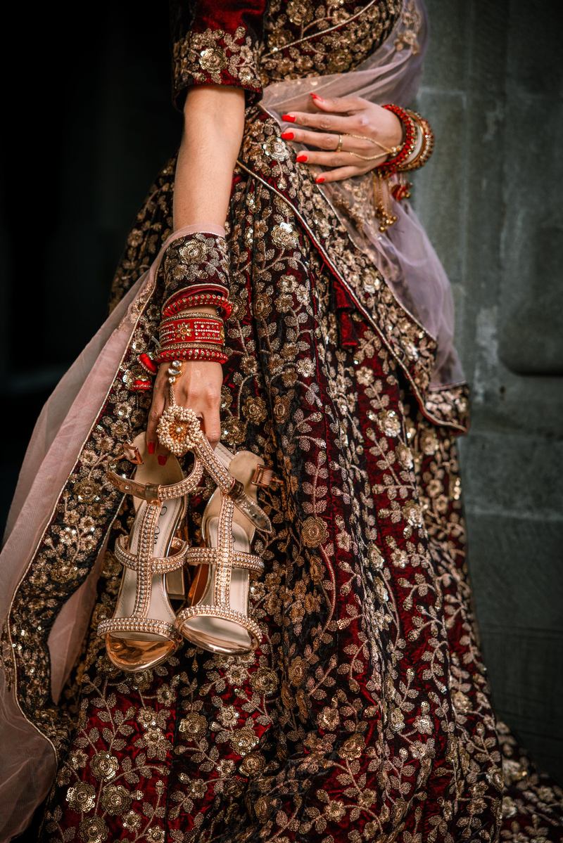 Bridal detail of jeweled gold heels, red bangles and a pearl hand ornament against an embroidered lehenga