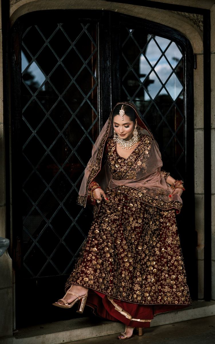 Bride stepping through an arched lattice doorway in a maroon embroidered lehenga and gold heels