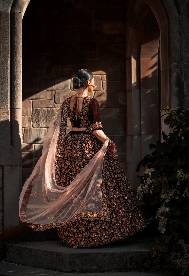 Bride seen from behind beneath a stone archway, her dupatta flowing in warm light