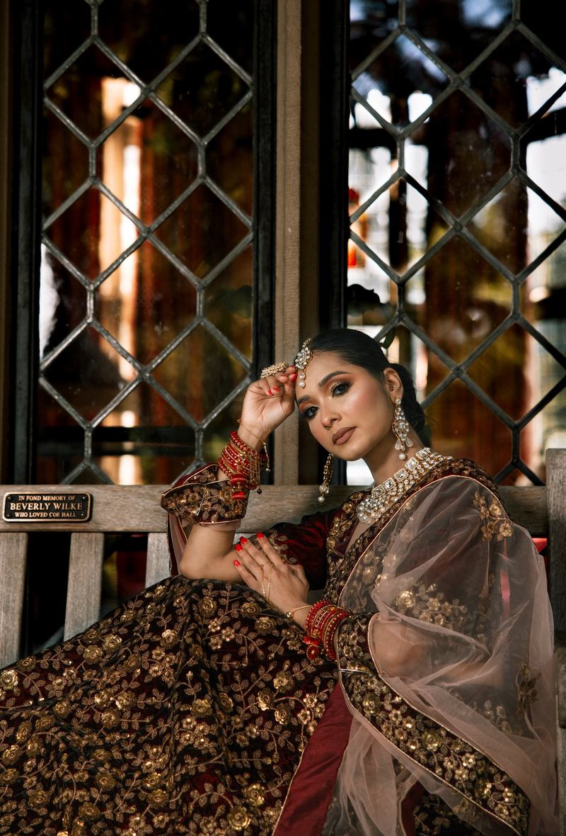 Bride seated on a wooden bench beside a leaded-glass window, hand resting at her maang tikka