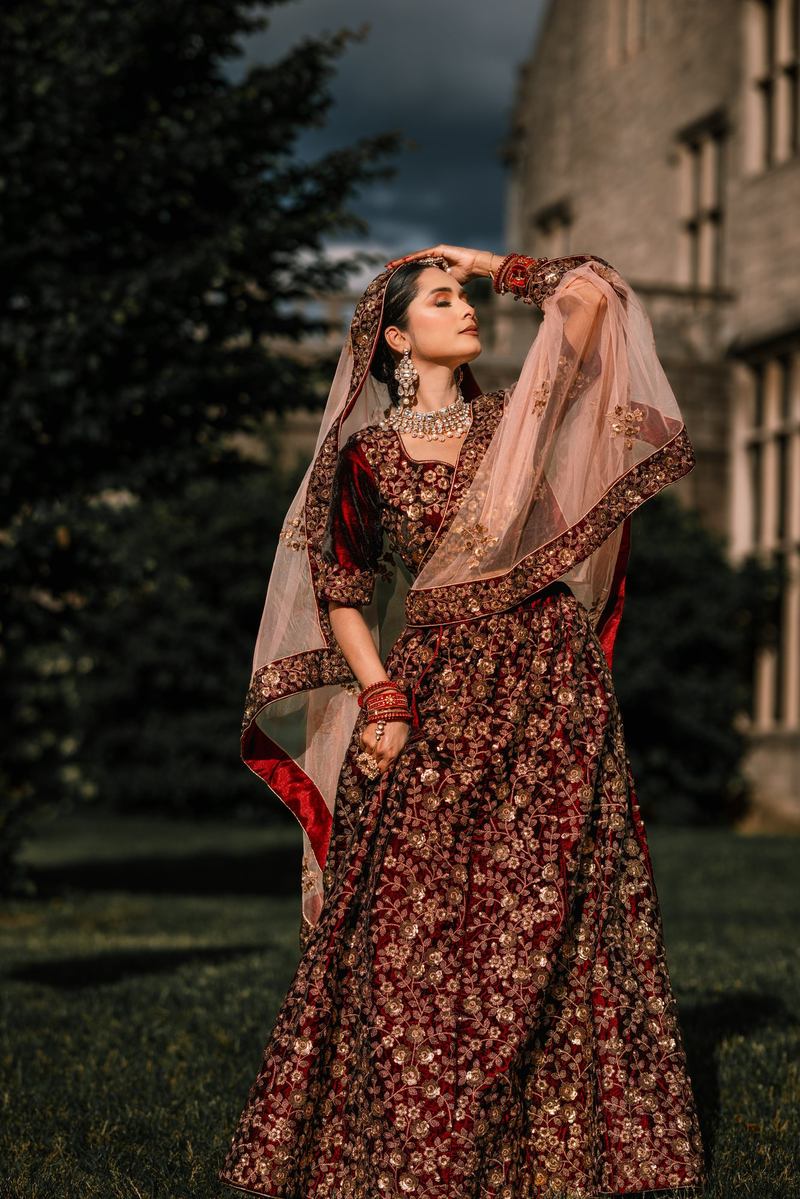 Bride in a red embroidered lehenga raising her hand to her dupatta on a lawn beneath a stormy sky