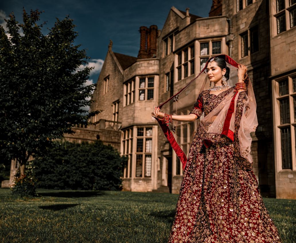 Bride lifting a sheer blush dupatta on the lawn of a stone Tudor mansion